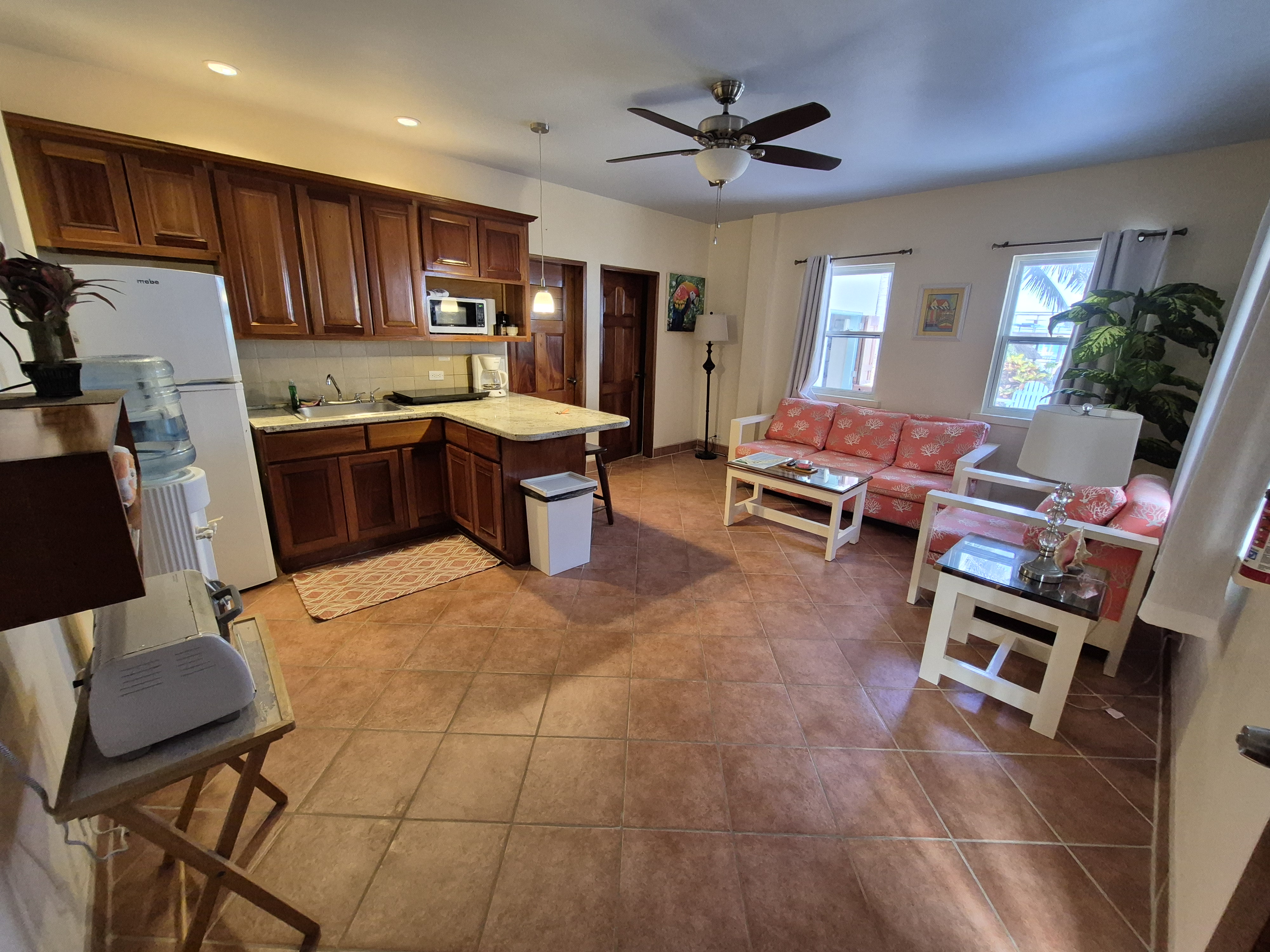 Indoor view of a kitchen and living area with wooden cabinets, white appliances, tiled floor, and a comfortable sitting area in the background.