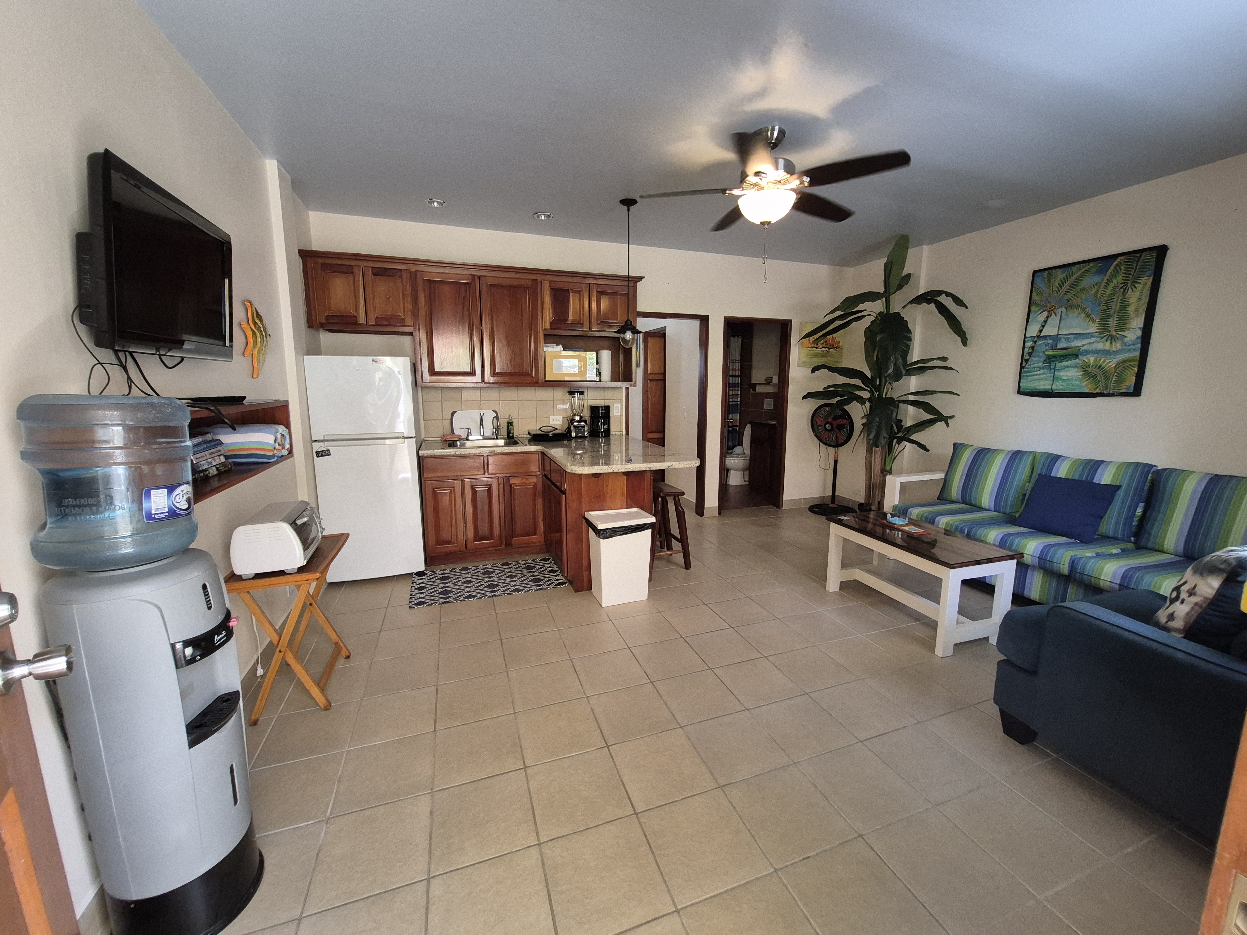 A seating area with couch, armchair, coffee table, two side tables with lamps, tv mounted on the wall and full kitchen.
