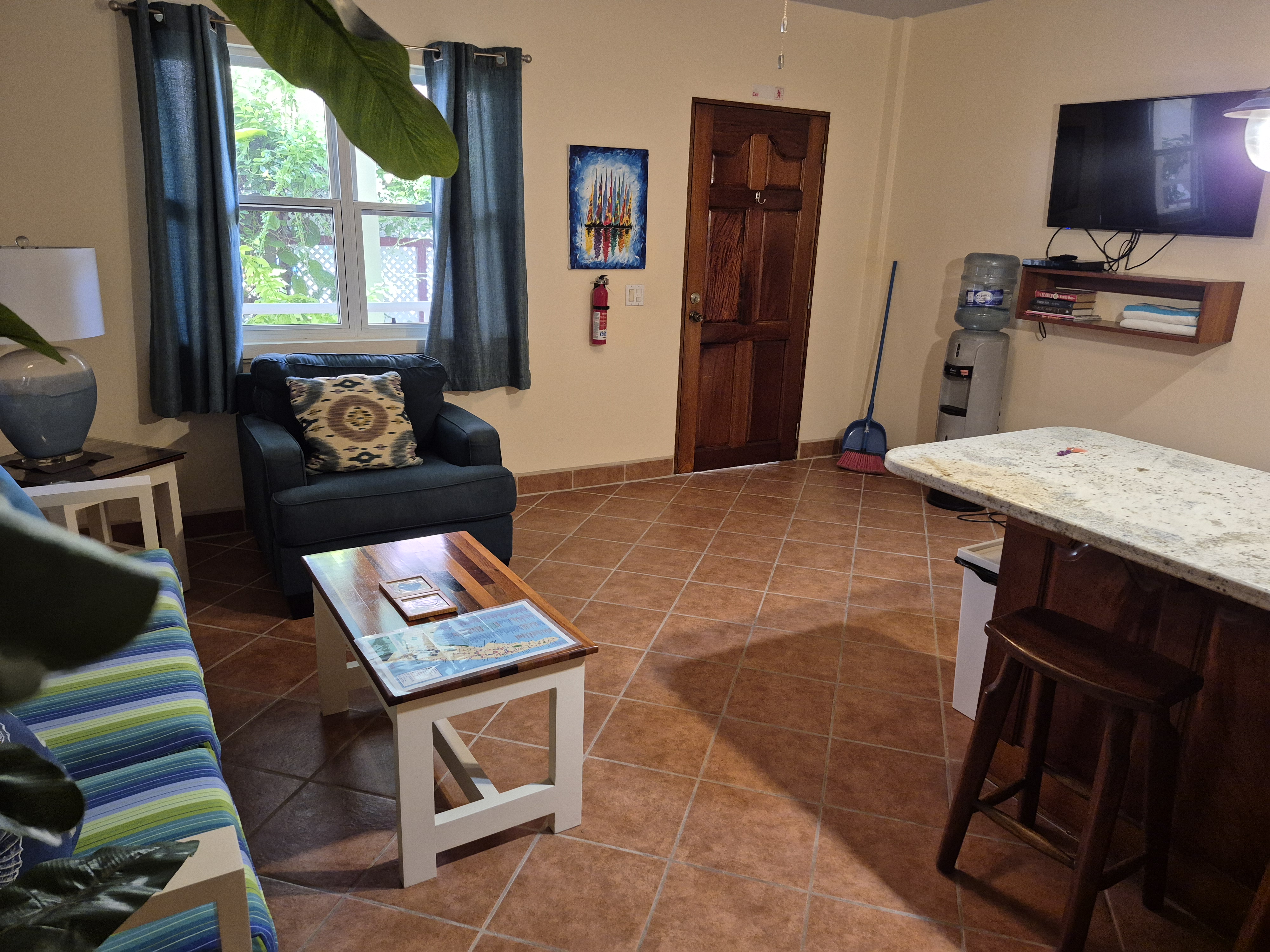 Living area with a blue armchair, striped sofa, white coffee table, and a breakfast bar with stools. A TV is mounted on the wall.