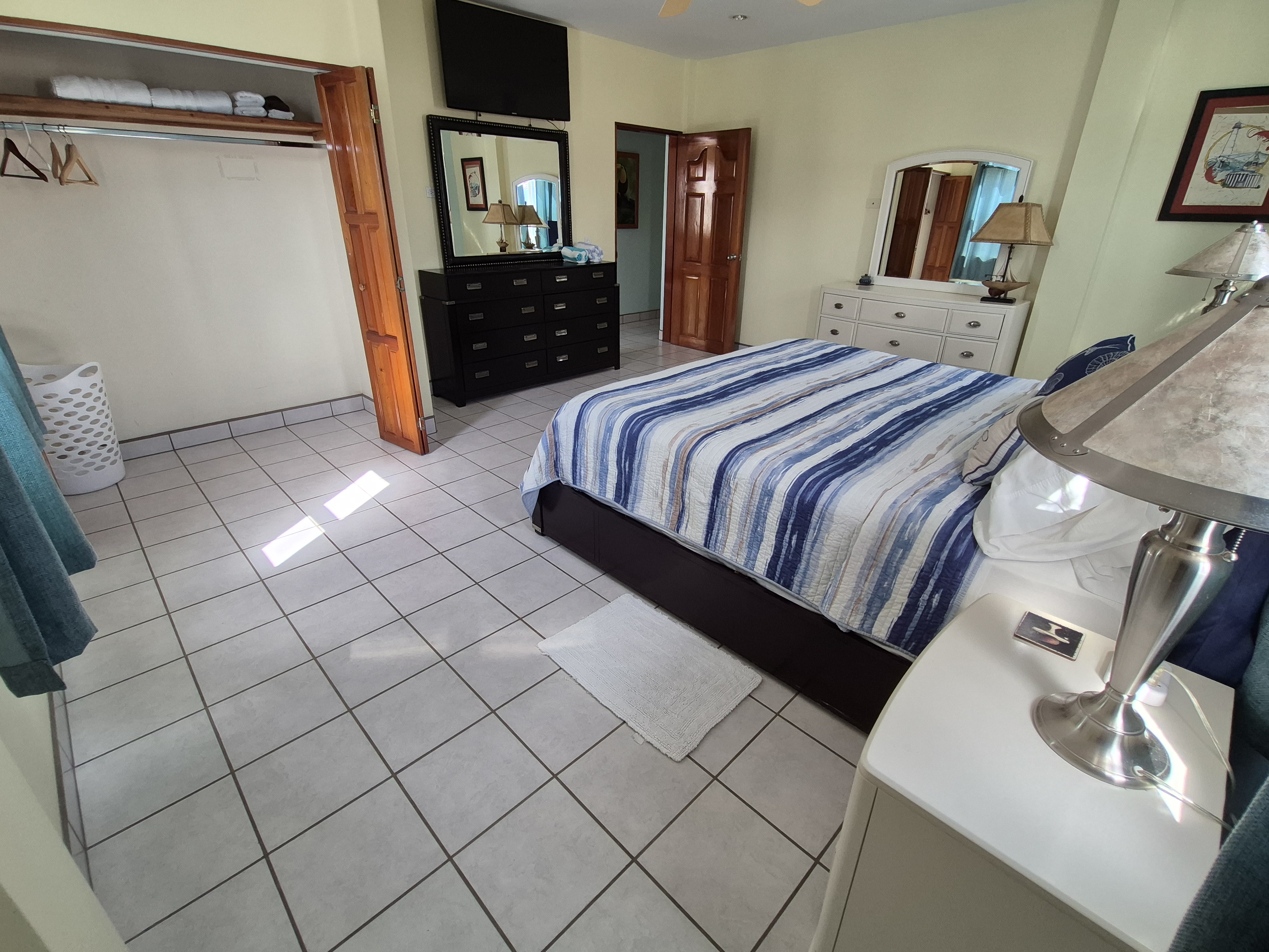 Queen bed with blue and white striped bedding, a nightstand with a lamp, and white tiled floors, light yellow walls, a dresser with mirror, and a flat-screen TV mounted on the wall.