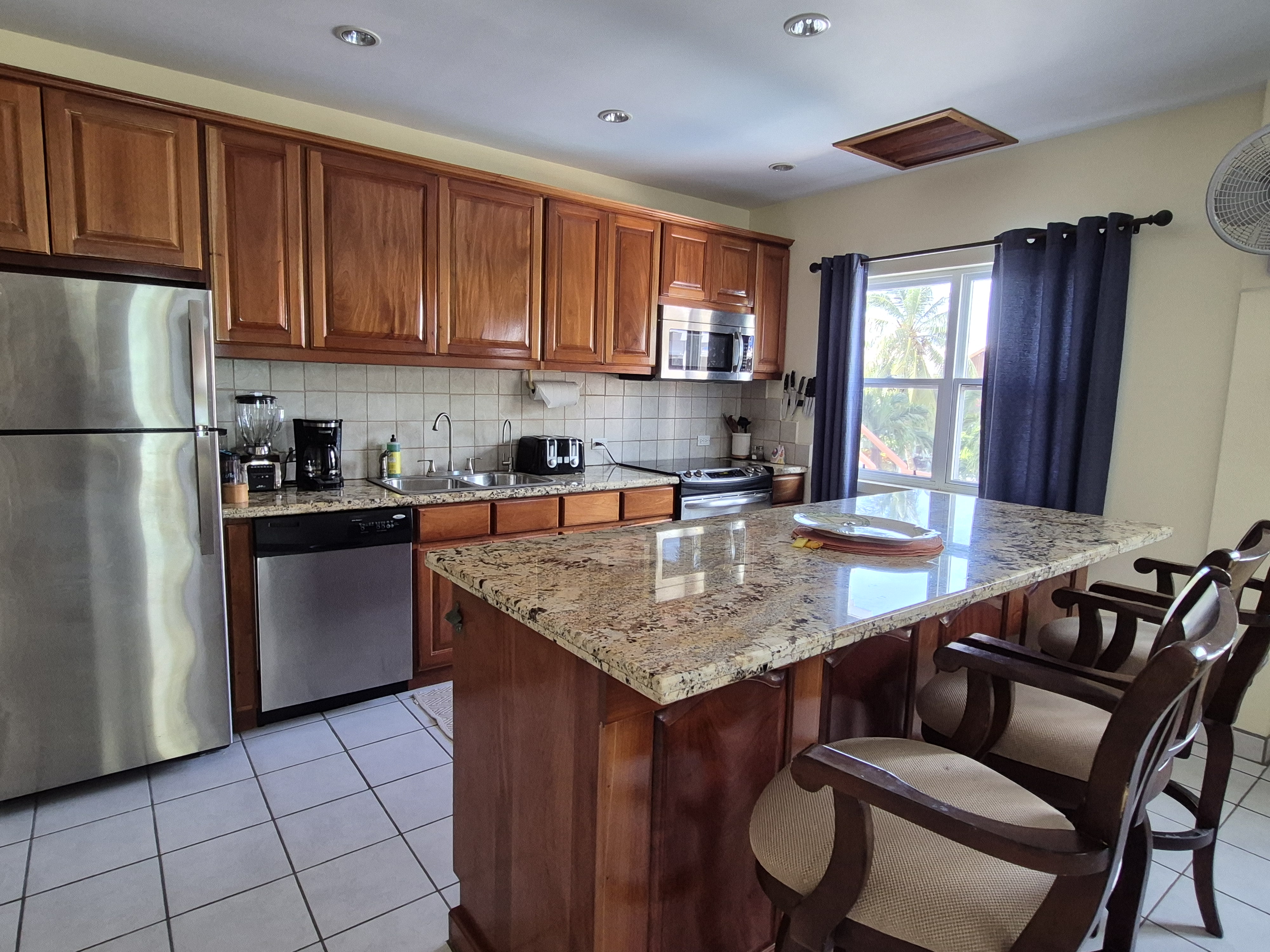 Kitchen with wooden cabinets, stainless steel appliances including a refrigerator, dishwasher, and microwave, a kitchen island with three bar stools, white tiled flooring, and a window with dark blue curtains.