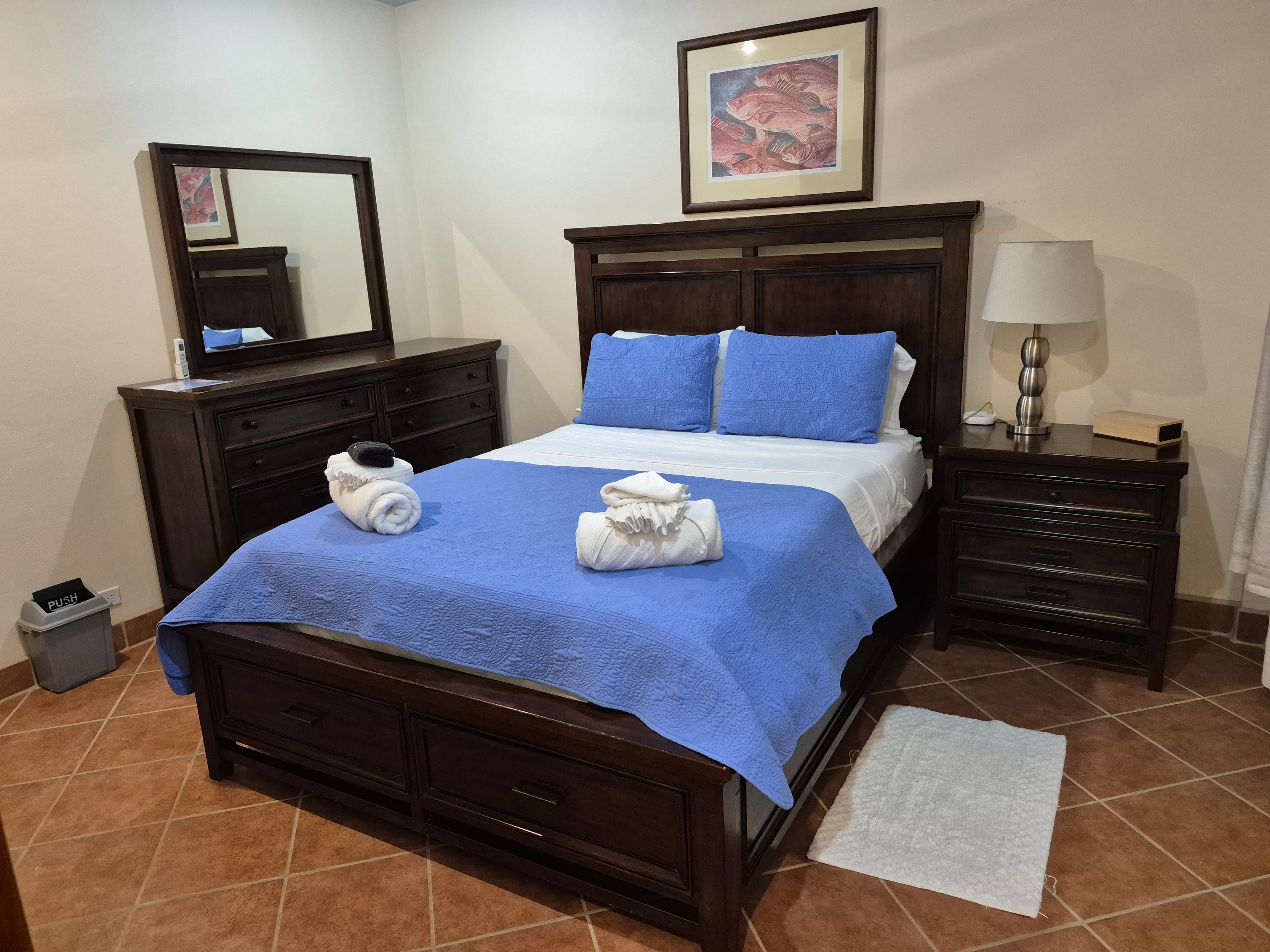 Ground-level bedroom with a dark wood queen-sized bed, dark wood nightstand and dresser, pale beige walls, and a brown tile floor.