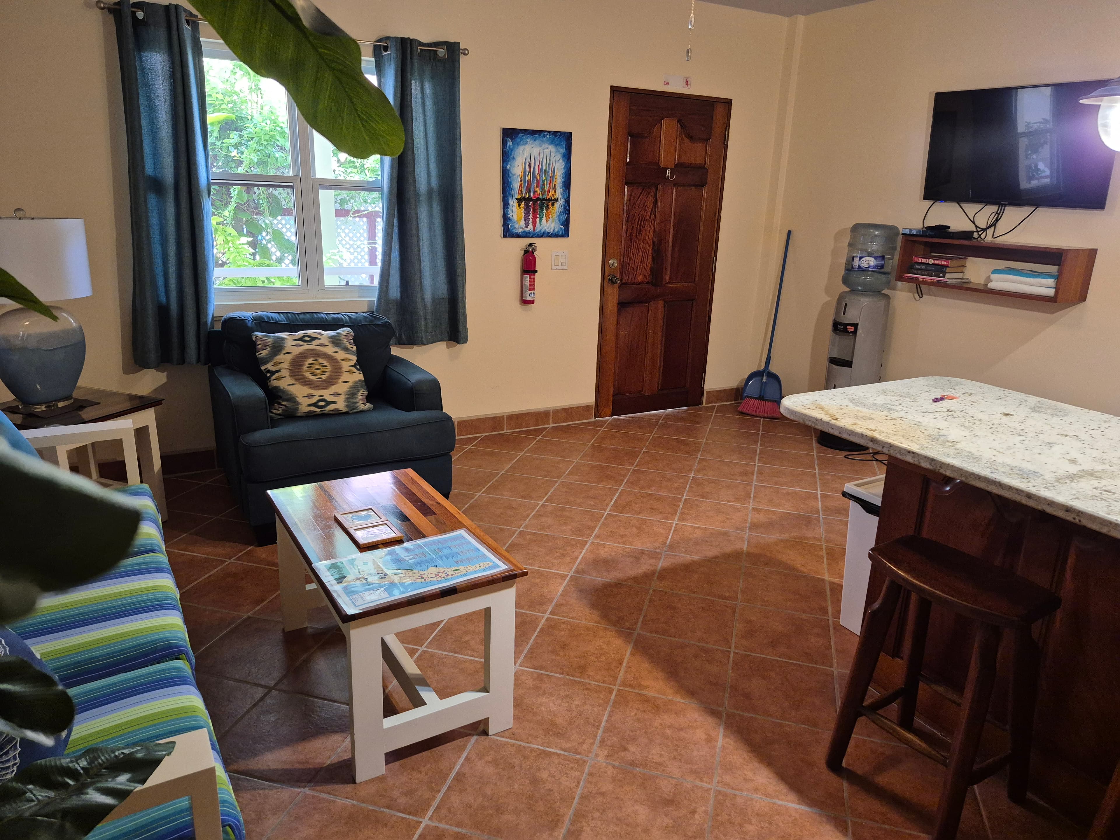 Living area with a blue armchair, striped sofa, white coffee table, and a breakfast bar with stools. A TV is mounted on the wall.