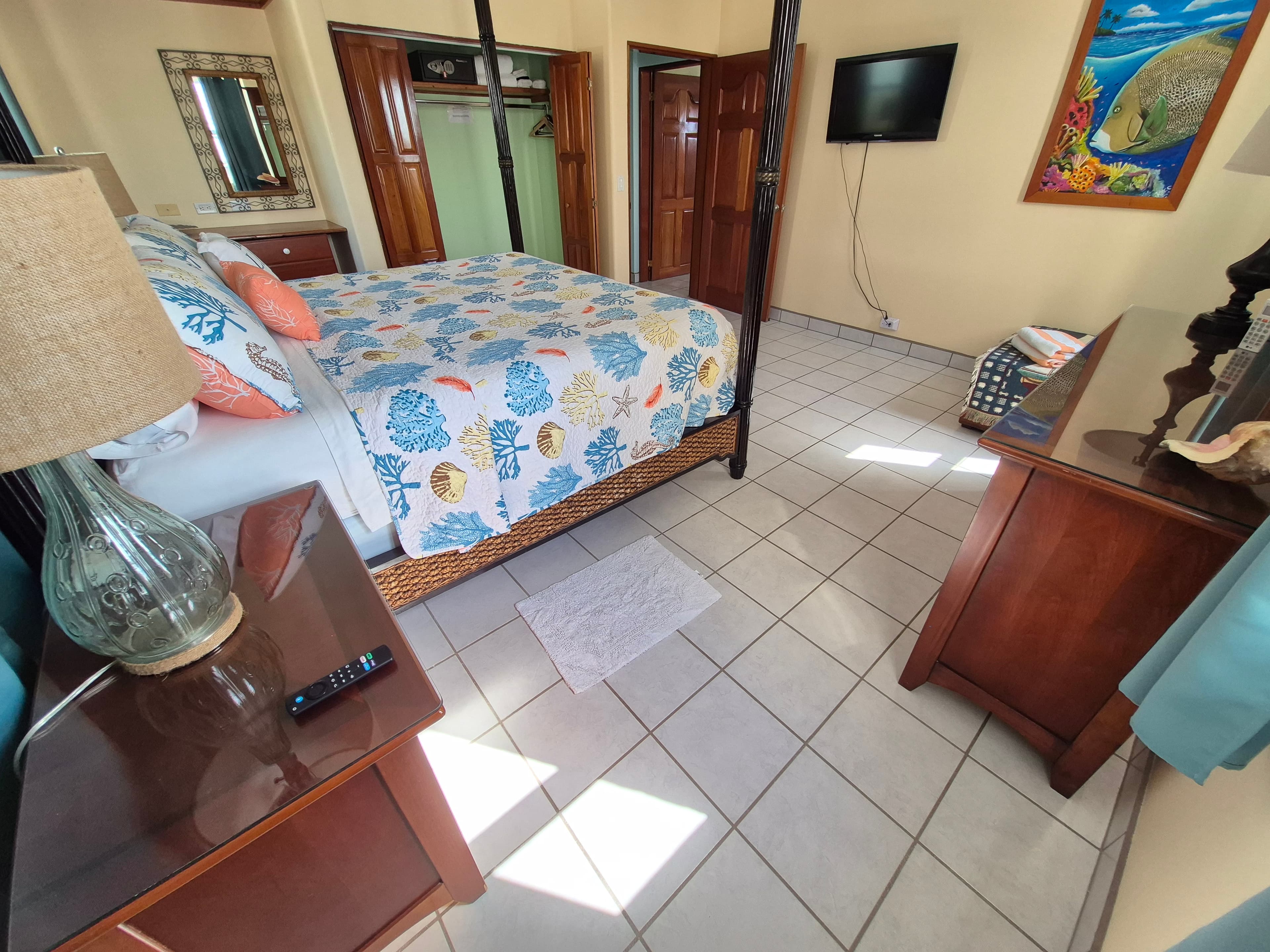 Bedroom with a four-poster bed featuring blue and white patterned bedding, dark wood furniture including a nightstand and dresser, tiled flooring, and a flat-screen TV mounted on the wall.