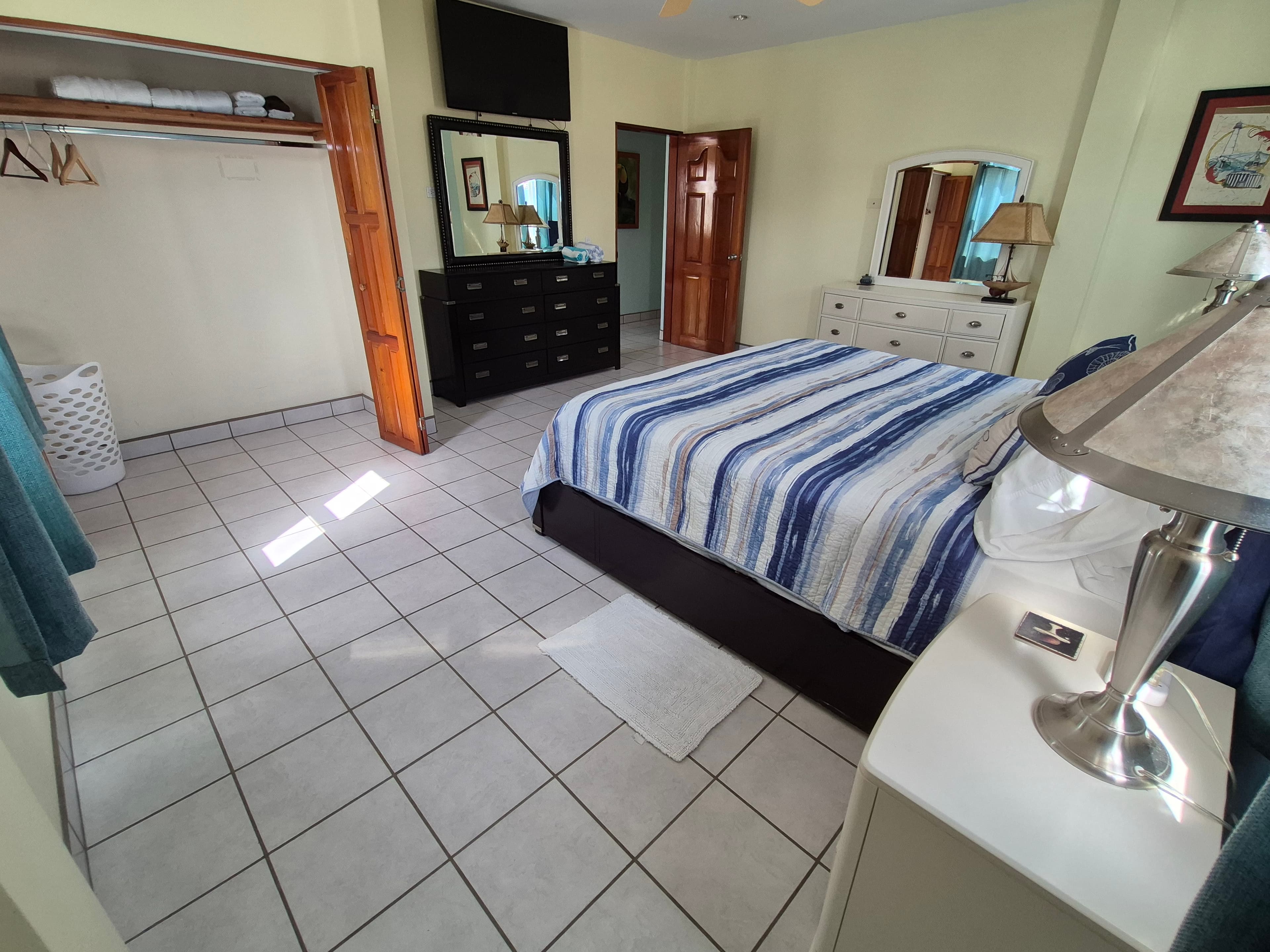Queen bed with blue and white striped bedding, a nightstand with a lamp, and white tiled floors, light yellow walls, a dresser with mirror, and a flat-screen TV mounted on the wall.