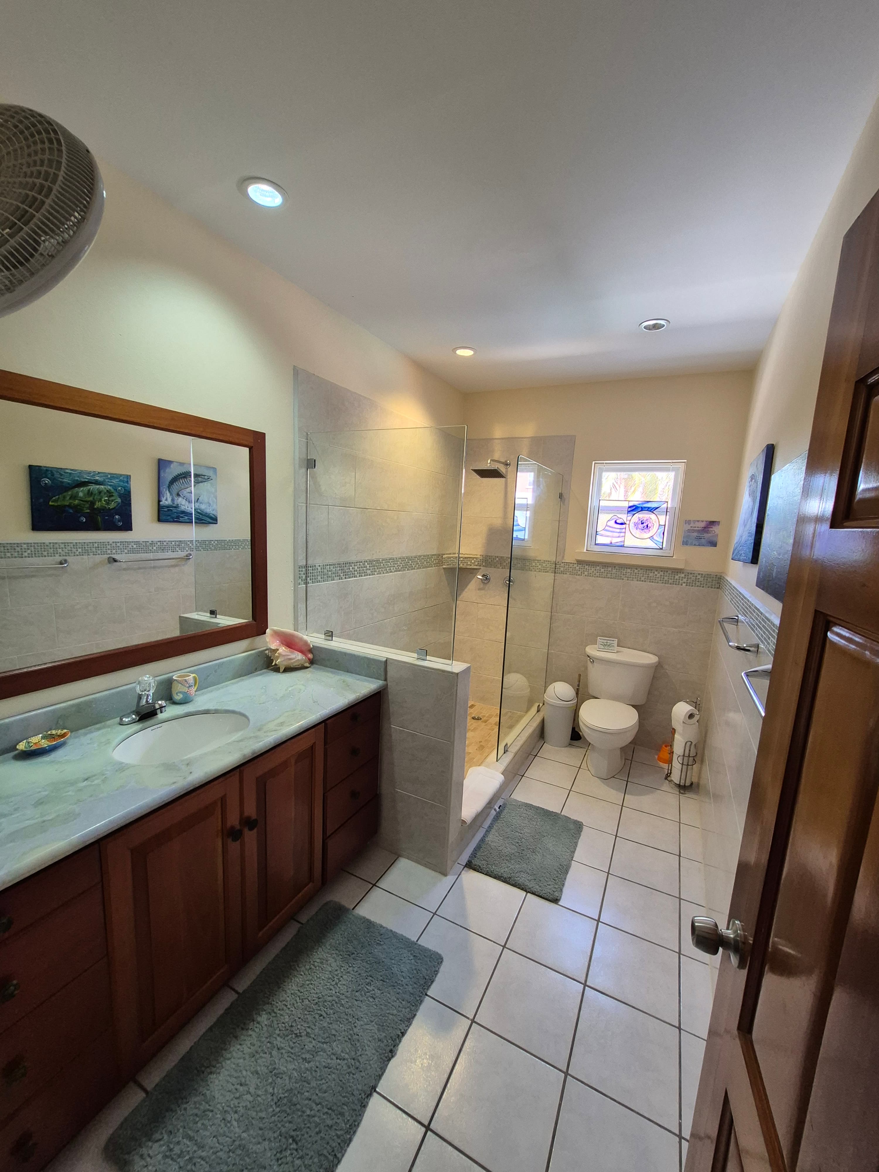 Spacious bathroom with a double sink vanity with wooden cabinets and a light countertop, a large rectangular mirror, a glass-enclosed shower, and a toilet.