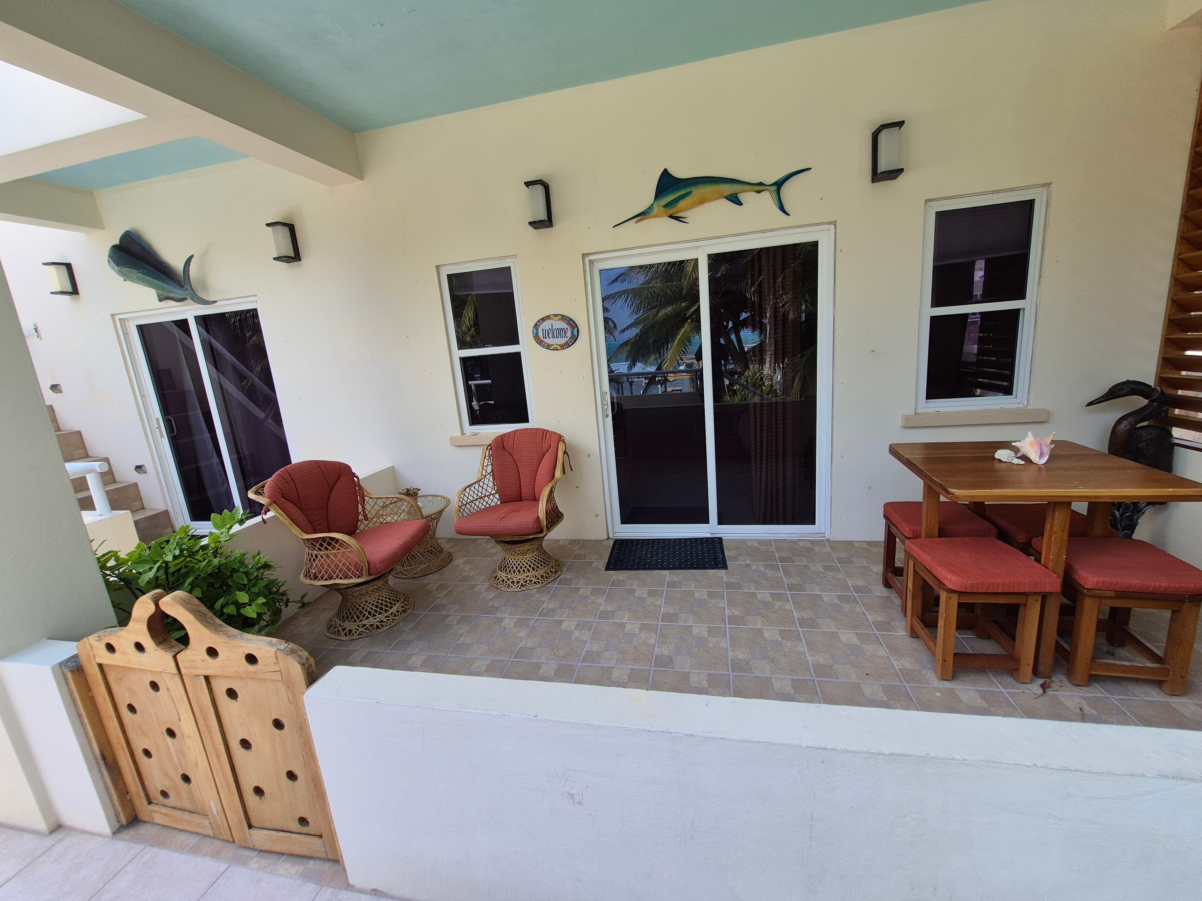Outdoor patio area with tiled flooring, light-colored walls, and a light blue ceiling. The patio features two red cushioned chairs, a wooden table with red cushioned stools, sliding glass doors, and several windows.
