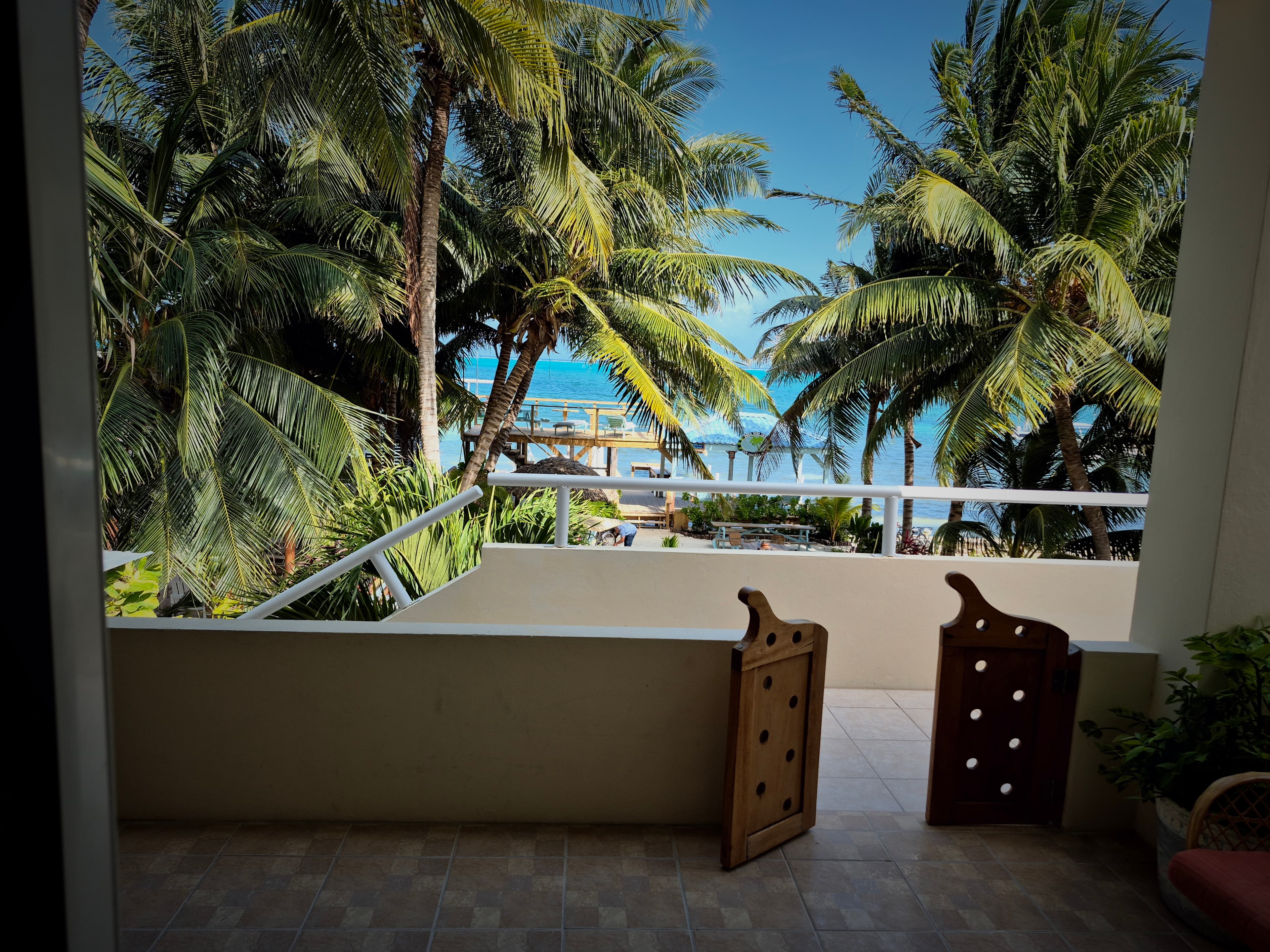 Balcony with tiled flooring and a white railing, overlooking a view of palm trees, the ocean, and a clear blue sky.