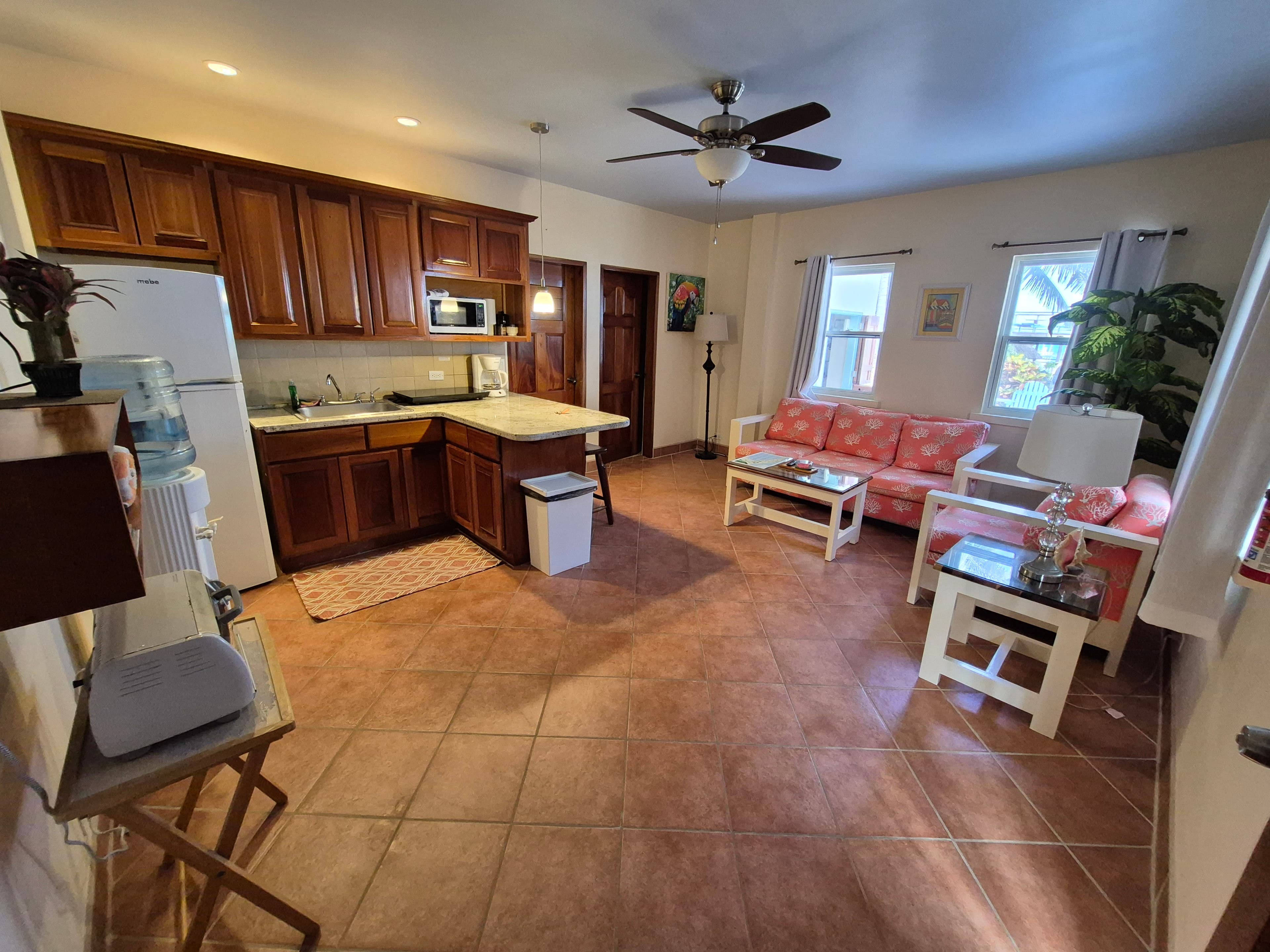 Indoor view of a kitchen and living area with wooden cabinets, white appliances, tiled floor, and a comfortable sitting area in the background.