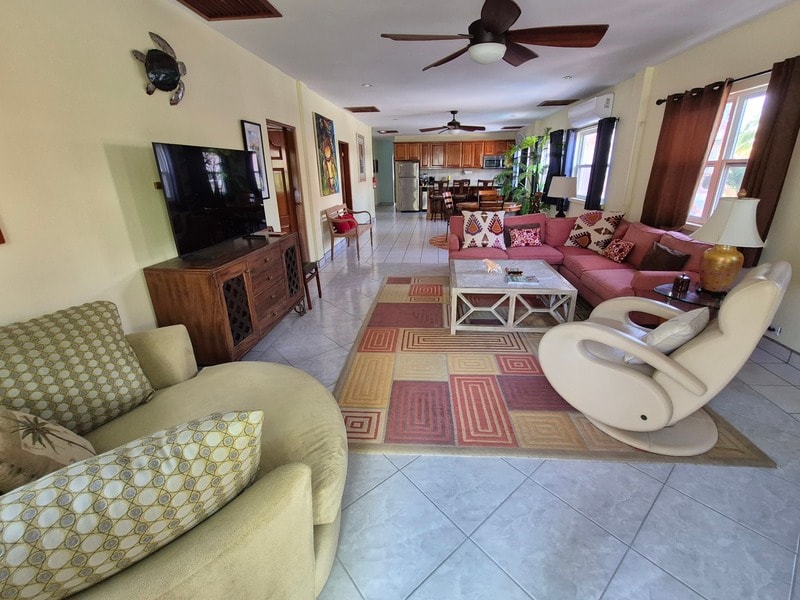 A spacious living room with modern furniture, a large TV, and a kitchen visible in the background.