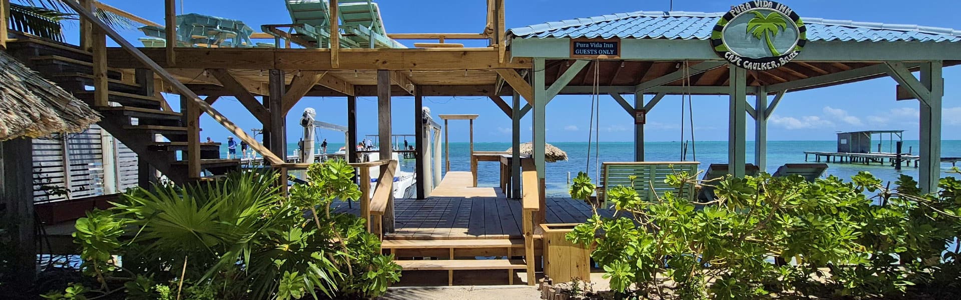 A wooden deck and stairs lead to the turquoise sea at Pura Vida Inn in Caye Caulker, Belize, surrounded by tropical greenery.