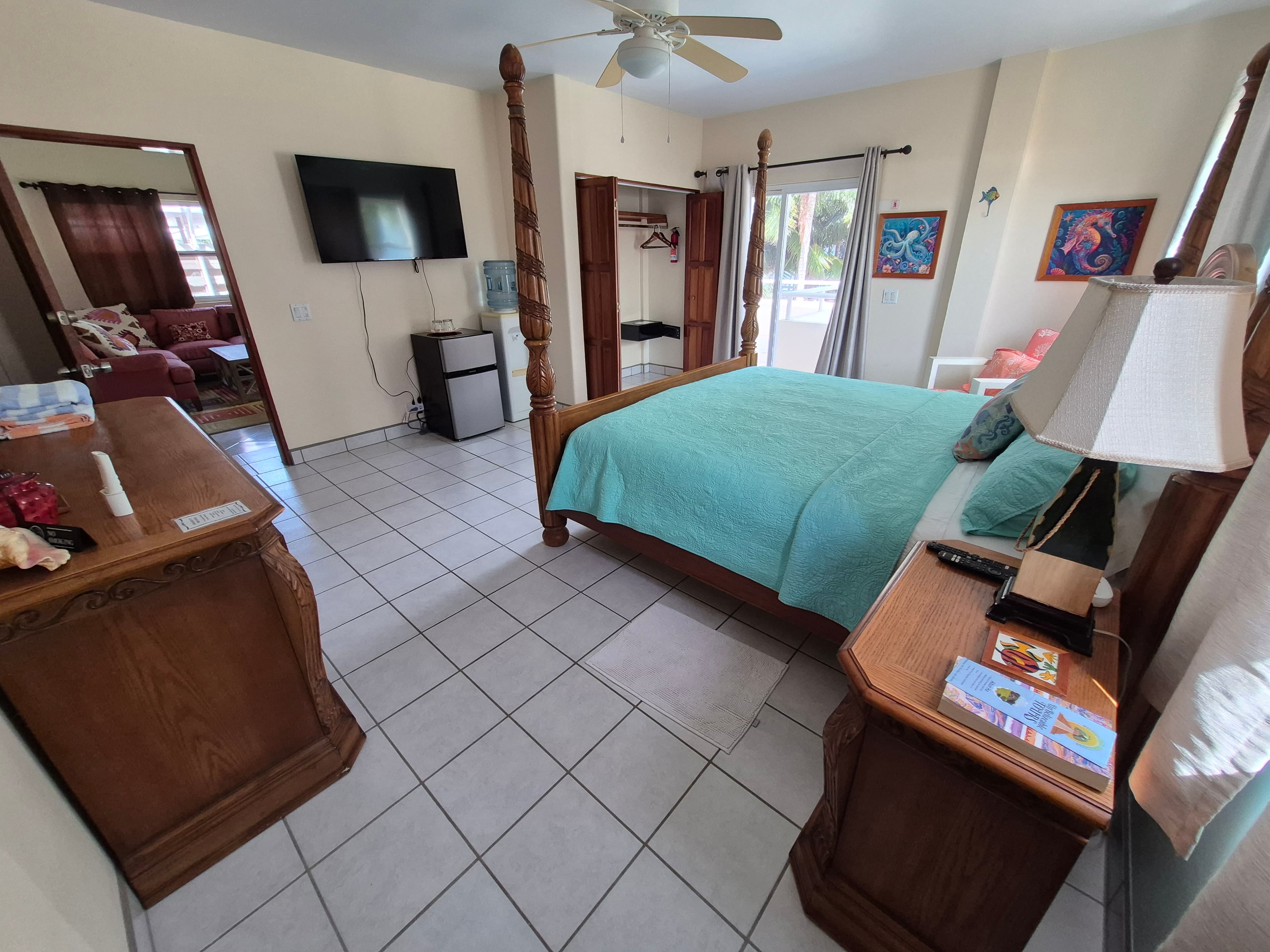 A bright bedroom featuring a queen-sized bed, wooden furniture, a television, and large windows opening to a patio.